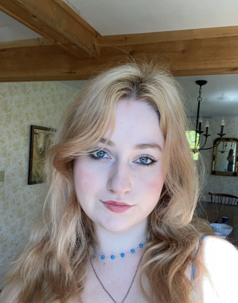 White girl with blue-grey eyes, wavy strawberry-blonde hair and very pink cheeks. She is smiling slightly with mouth closed, lips upturned, and wearing a blue beaded necklace, and you can see a bit of her matching blue spaghetti strap dress popping out from under her hair. 