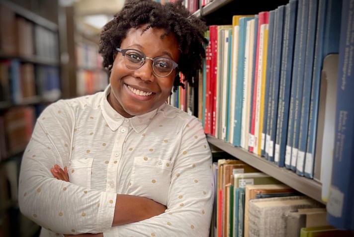 Dr. Bailey, a Black androgynous person with short natural hair, wearing glasses a white shirt, smiling and leaning against a library bookshelf.