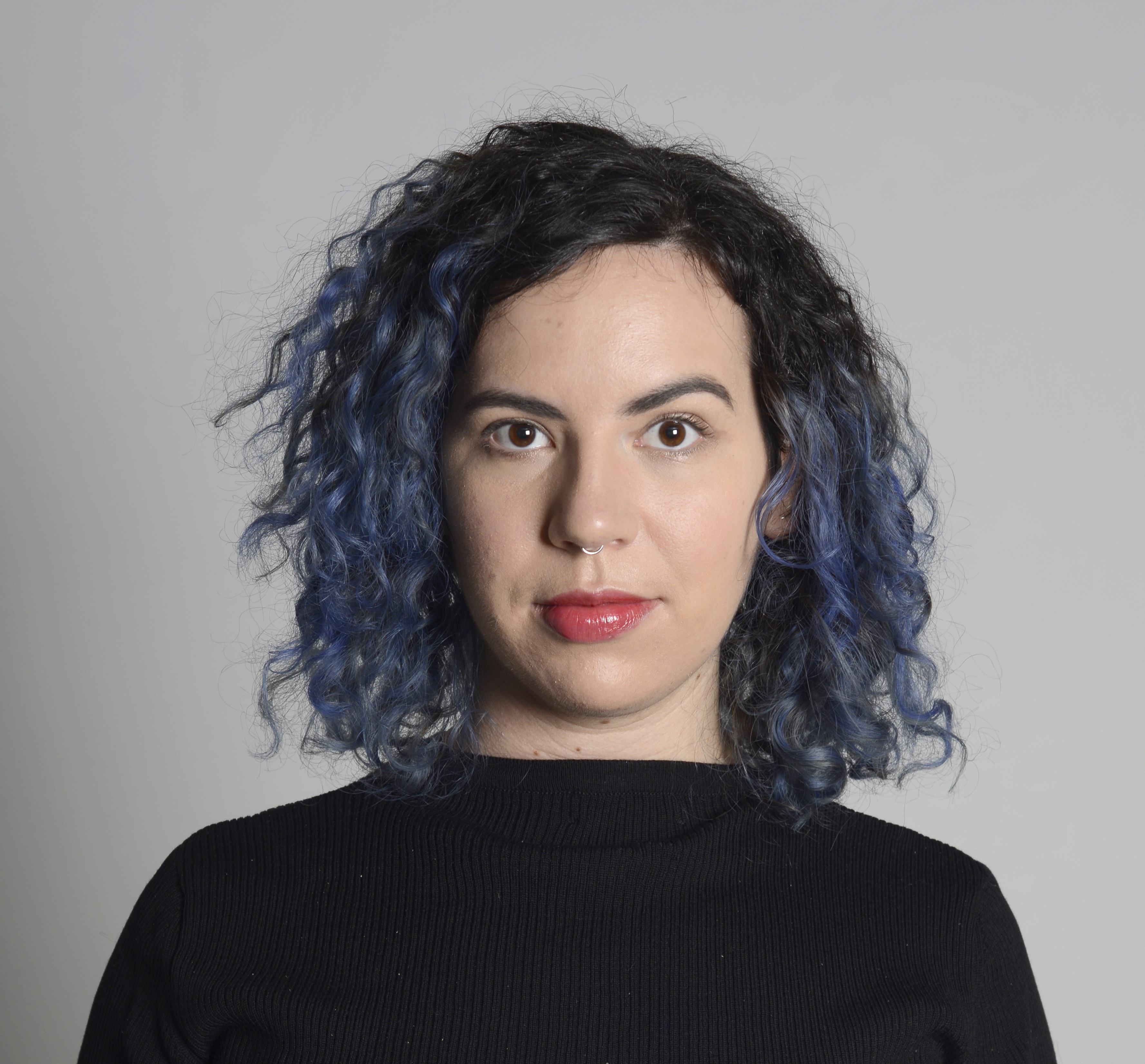 A headshot of a white femme-presenting person with shoulder length curly black and blue hair, wearing a black shirt, standing against a light gray background.