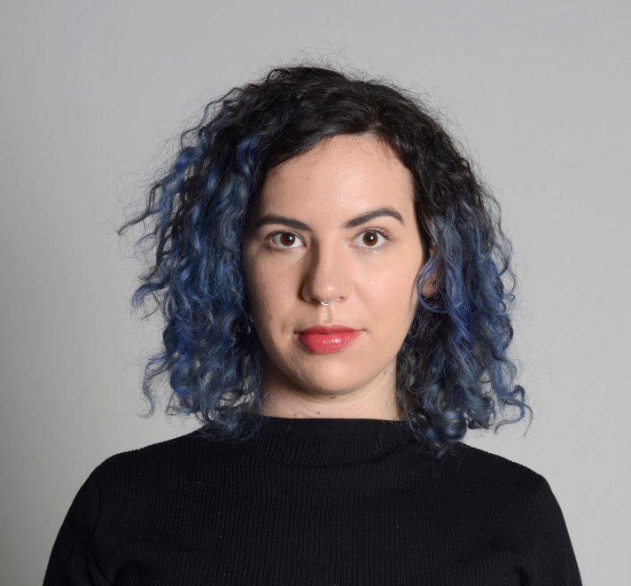 A headshot of a white femme-presenting person with shoulder length curly black and blue hair, wearing a black shirt, standing against a light gray background.