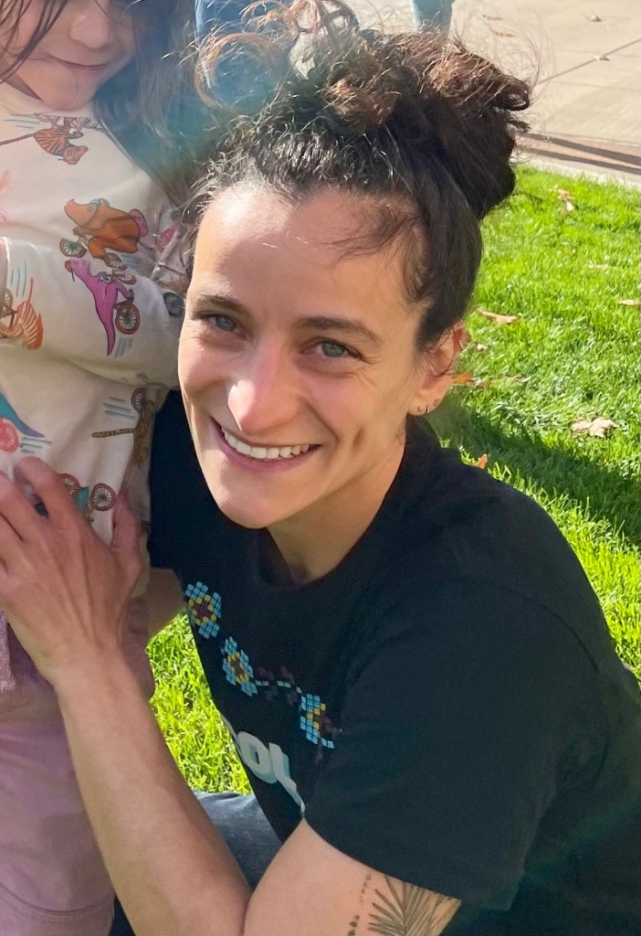 A white woman smiles at the camera. She has blue eyes and dark brown hair tied up in a high bun, and she's wearing a black t-shirt with illegible blue and white text. She's kneeling in a grassy field next to a child, who is cut out of the frame, save for a white dinosaur-printed shirt.