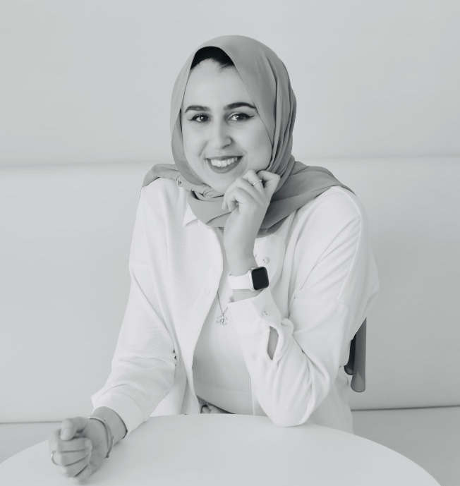 A black and white headshot of Jumanah Abbas wearing a white shirt and a grey head scarf and partially smiling at the camera. in an empty coffee shop.