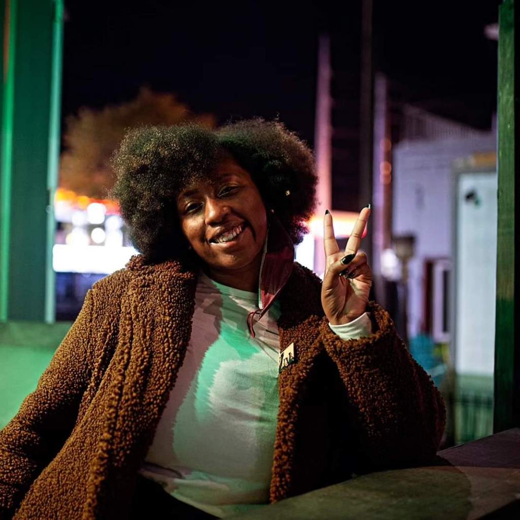 Photo of a brown skinned person leaning against a bar throwing up a peace signed with stiletto black and green nails. They are wearing a white shirt, a brown teddy bear coat with a yellow button and “power” in black letters in the shape of fists. This person has an afro with dark brown highlights, a side part and a singular twist with a gold clasp at the end. They are wearing a partially worn burgundy kn94 mask, taken off to reveal a smile.