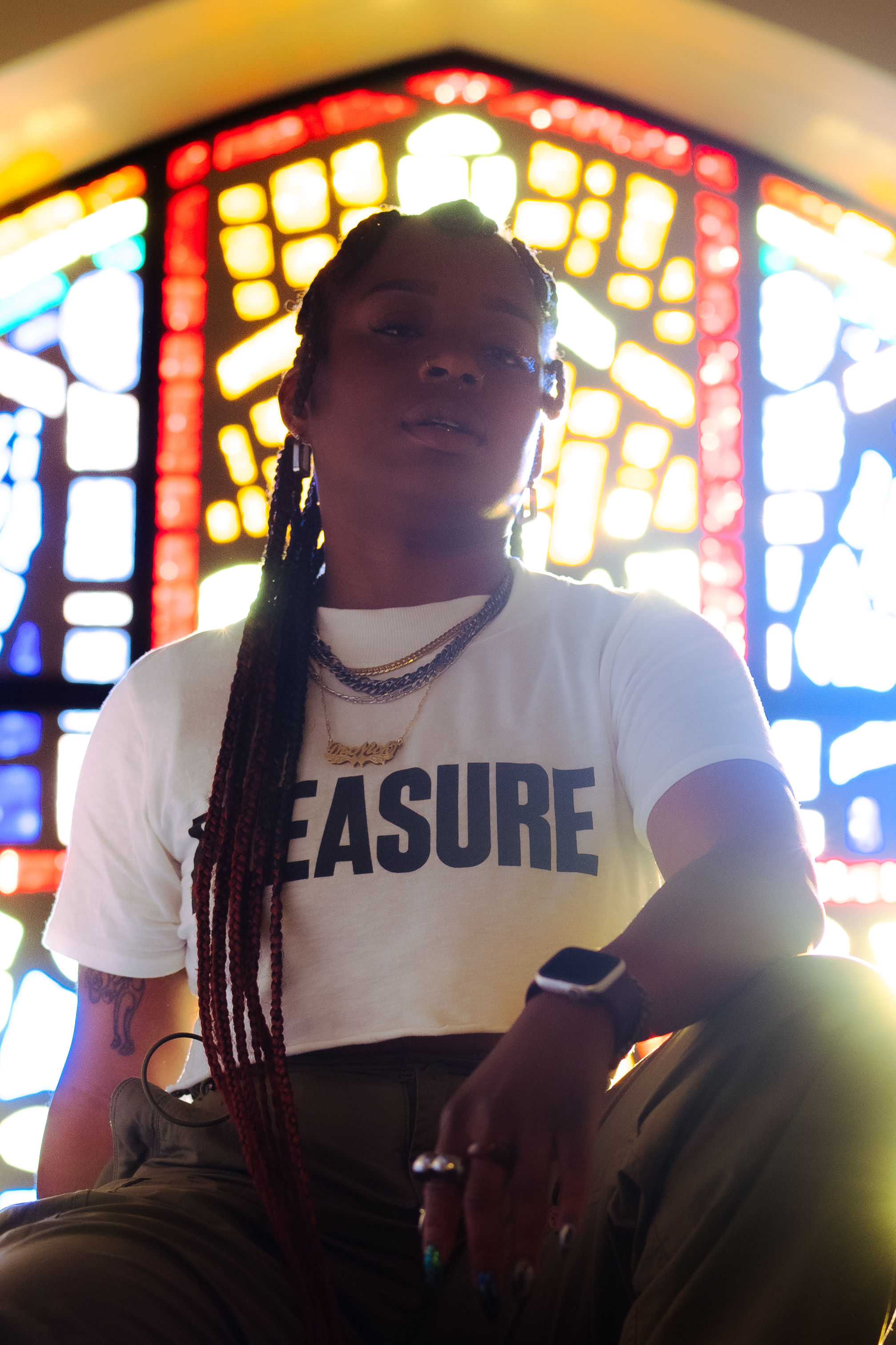 This image features a brown-skinned, non-binary person standing confidently in front of a vibrant stained-glass window. The lighting from the window casts a warm, almost halo-like glow behind them. They have long braids and are wearing a white t-shirt that prominently reads "PLEASURE" in bold black letters. The person has multiple necklaces, including one with a nameplate that says “dr. nick.” They are also wearing a smart watch and various rings, adding a stylish touch to their look. Their posture exudes confidence, and they appear calm and grounded. The stained-glass window in the background adds a spiritual, almost ethereal element to the photo. The overall vibe is both powerful and reflective, merging personal style with a sense of deep meaning.