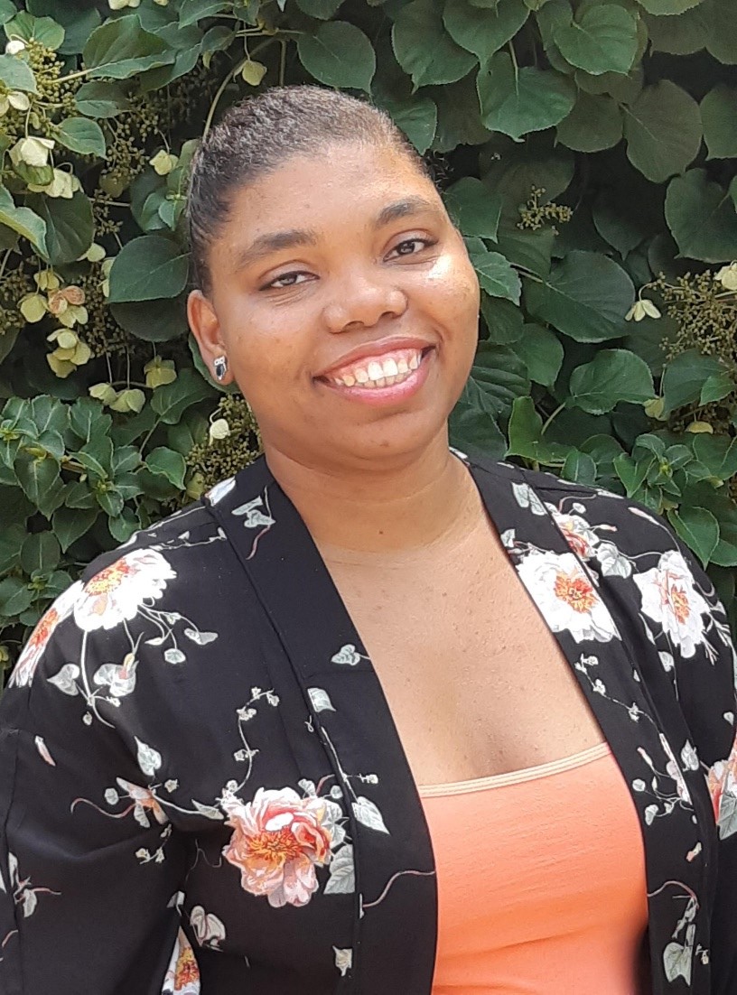 Photo of a brown skinned woman in front of a flowering plant. She is wearing a multicoloured floral shirt with a peach undershirt. She is facing the camera and smiling.