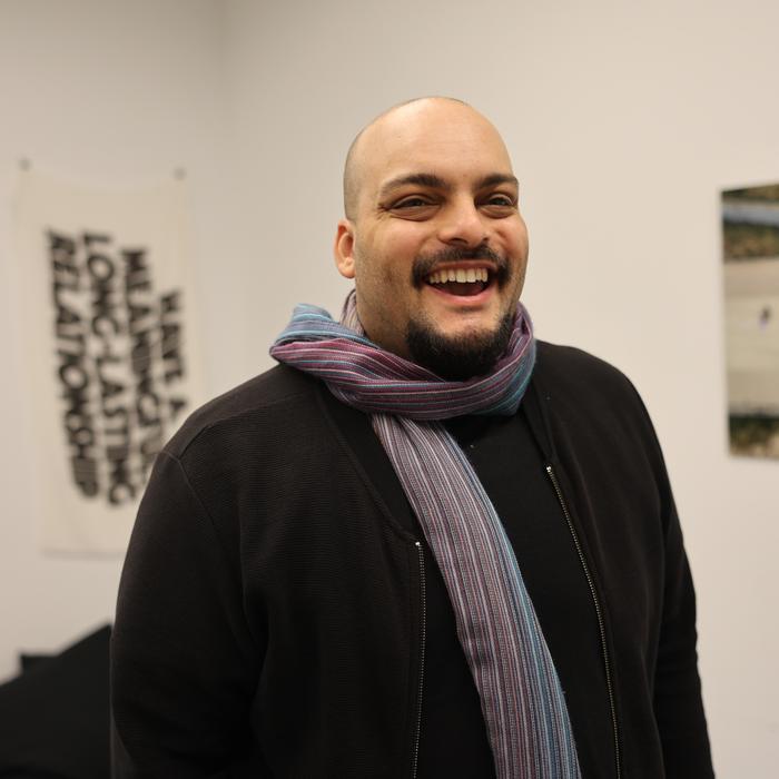 Jeff stands in a gallery, and smiles at the camera. He is wearing a purple-blue-grey scarf and a black shirt.