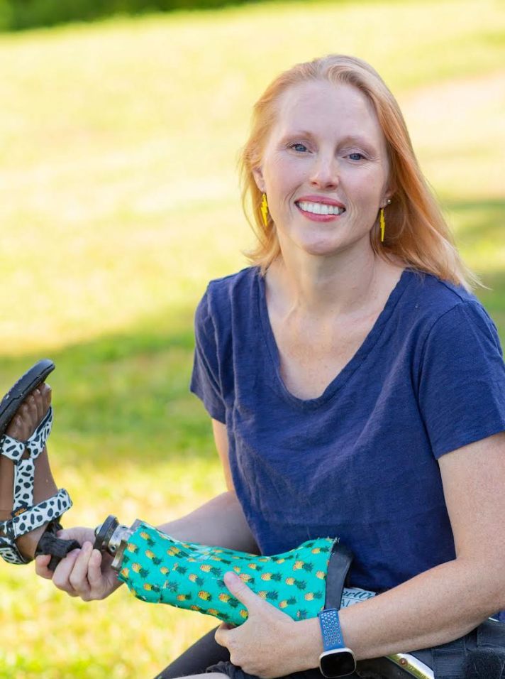 Ashley Shew, a white woman with reddish hair holds a leg prosthesis and smiles at the camera, outdoors on a pretty day.