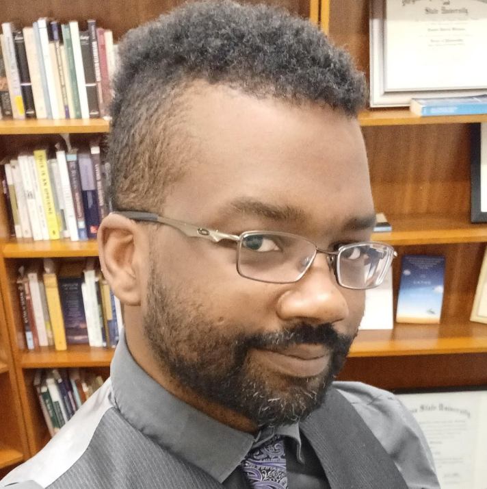 Self-portrait of a smiling Black man with natural hair shaved on the sides and long in the center, a beard, and grey square-frame glasses, wearing a silver-grey pinstriped waistcoat and a dark grey shirt with a purple paisley tie, and black jeans; bookshelves filled with books and framed degrees are visible in the background