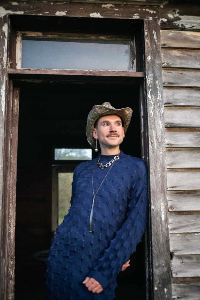 kevin stands in the doorway of a brown, wooden building, wearing a cowboy hat, long blue textured tunic, silver necklaces, and a dangling earring. 