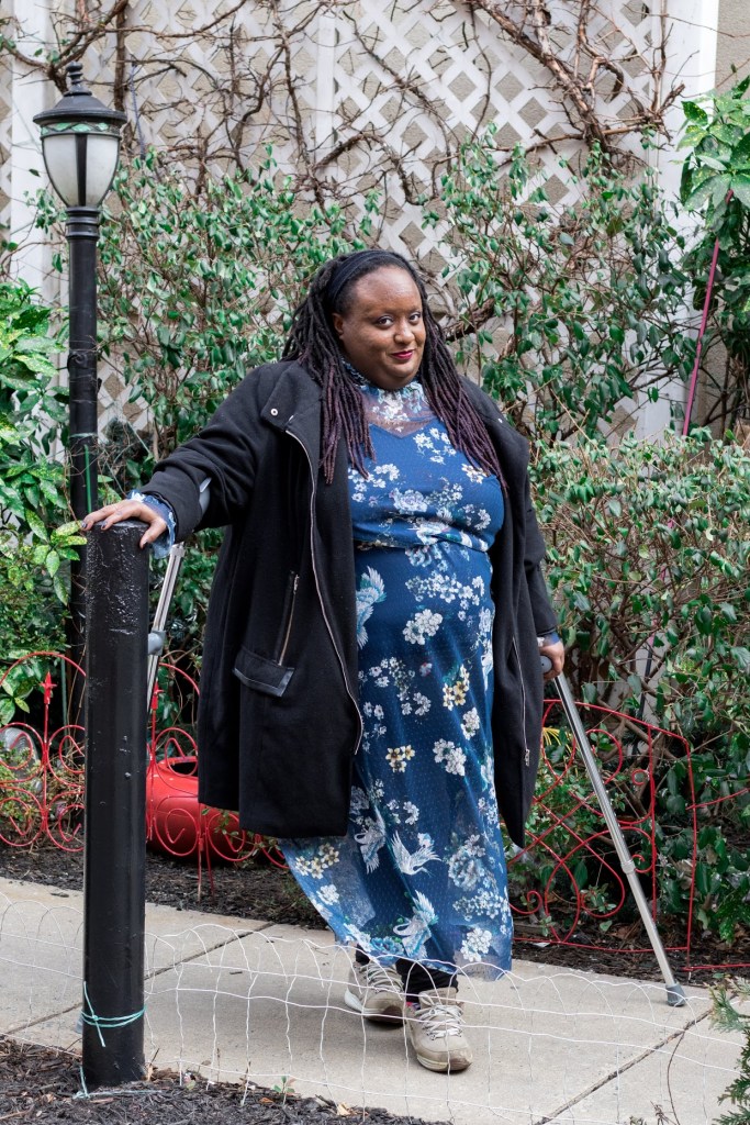 Imani Barbarin in a blue floral dress, black jacket, and sneakers, stands outside on a sidewalk, crutches on either side.