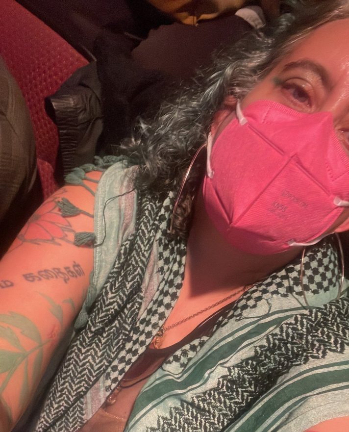 Leah wears a pink mask and a black, white, and green kuffiyeh, holding the camera up at an angle from above. Their arms are tattooed and they have black and white hair.