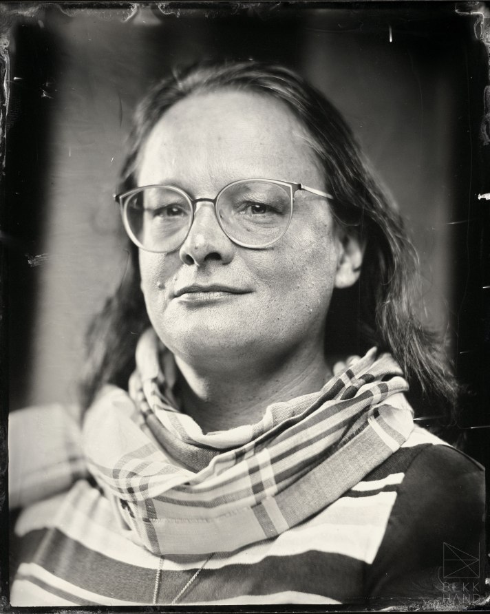 A grainy black and white, tintype image of Bess Williamson, a white middle-aged woman with dark hair and glasses, wearing a mix of stripes and plaids.
