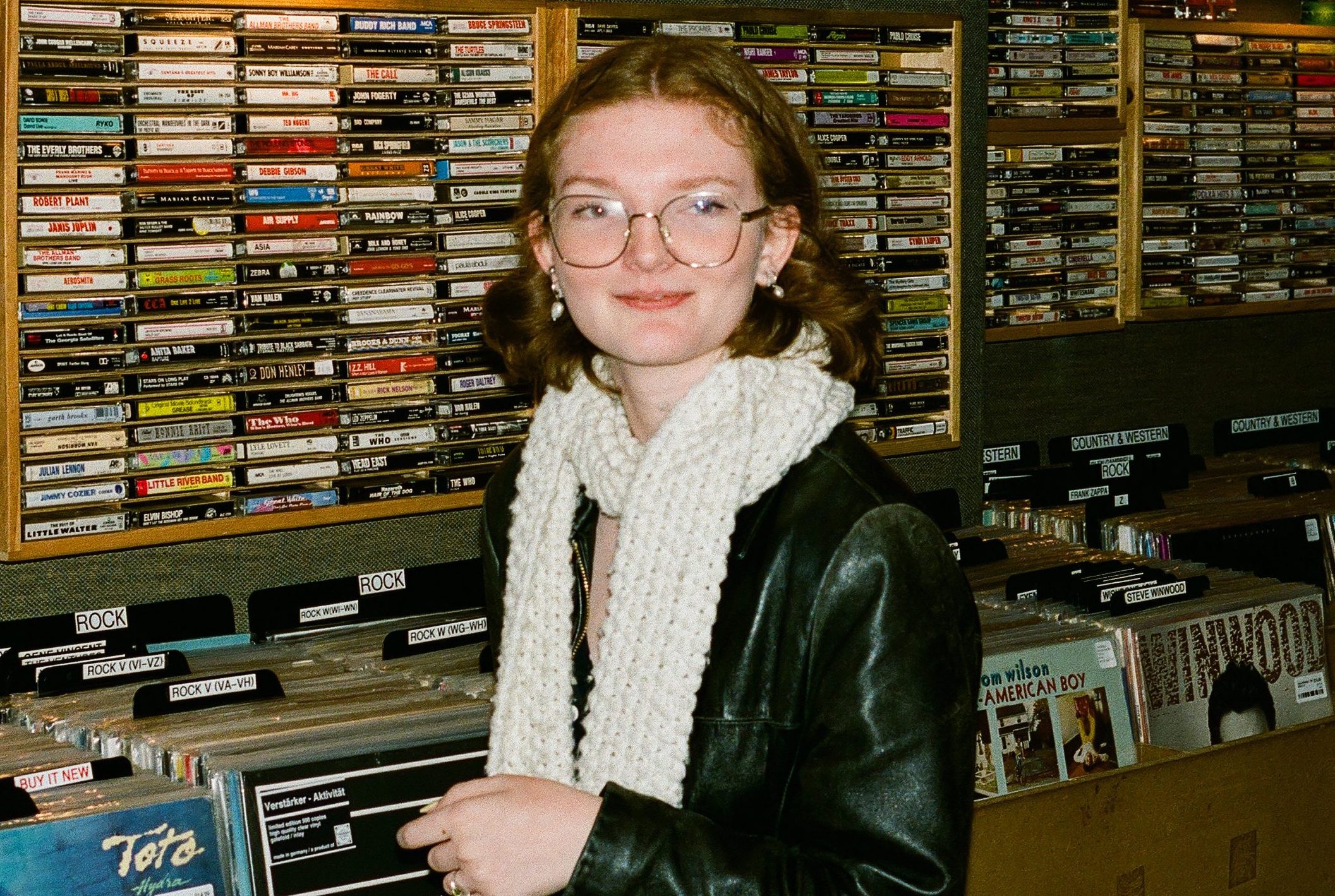 Film portrait of Katie, a white femme with a curly auburn bob. She's bespectacled in gold octagonal frames, wrapped in a big white scarf and a leather jacket. She grins awkwardly in front of a wall of cassette tapes in a record store basement.